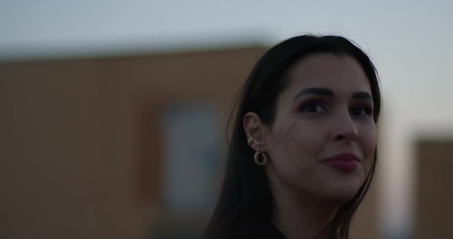 Woman with dark hair earrings and black overcoat happily looks around on top of building at sunset with city skyline in the background