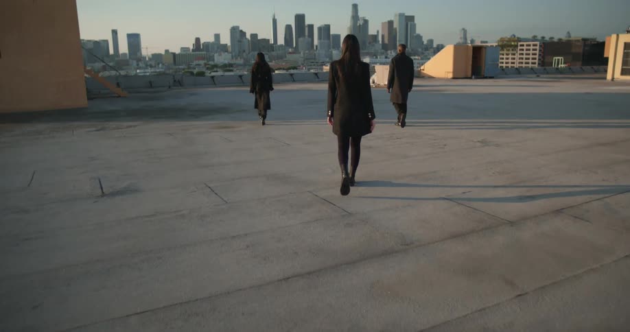 Three women dressed in black walk in slow motion in a triangle formation into the city