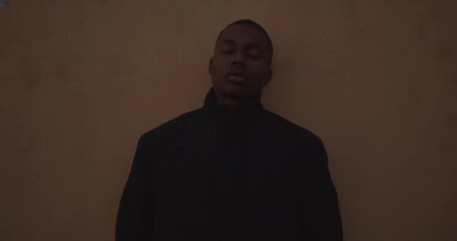 A man standing against a wall with his eyes closed wearing a black jacket