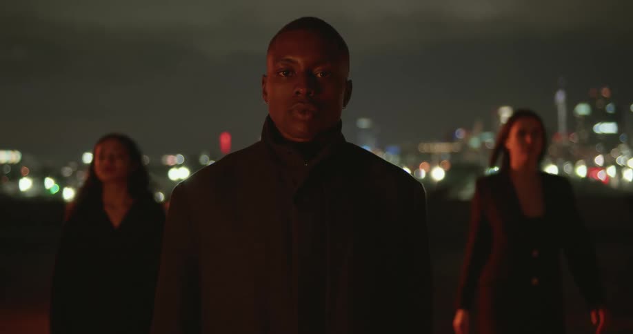 Man in black coat and women wearing black coat and blazer jacket walk on top of the building with city skyline in the background at night