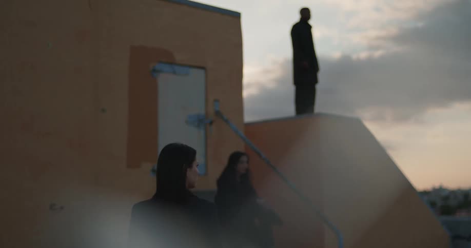 A man standing on a structure on the rooftop of a building at dawn while two women are watcing him one of the women is sitting on a flight of stairs waiting