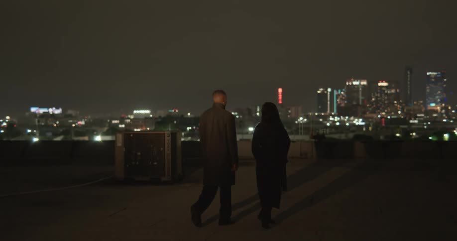 Woman walks across building roof lit by moving spotlight at night with skyline in the distance
