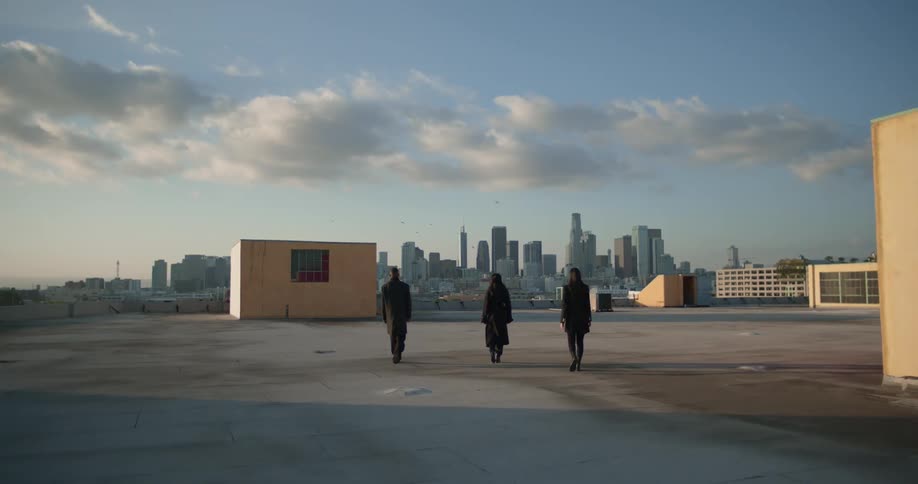 Three people wearing dark coats walk in unison across parking lot toward skyline under a cloudy sky