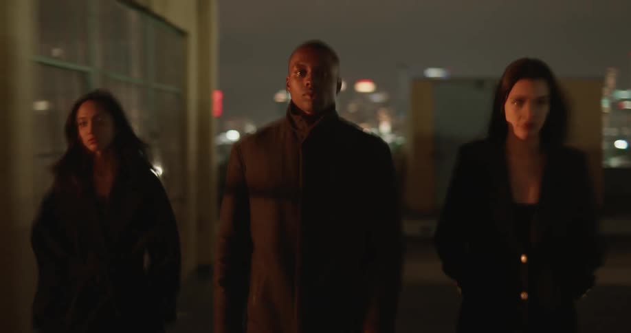 Three people walk over building roof top with city lights in the background at night