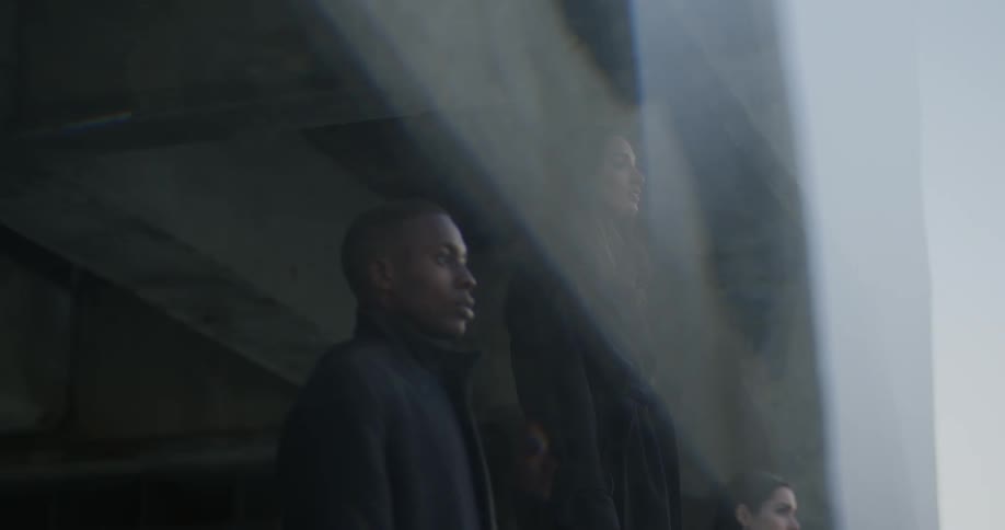 Man in black coat and women wearing black coat and blazer jacket stare out of large window of a building