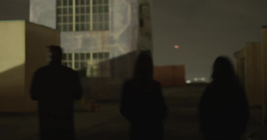 Two women and a man all wearing black are shown sped up walking near home buildings while a red lights flashes onto them from somewhere off camera