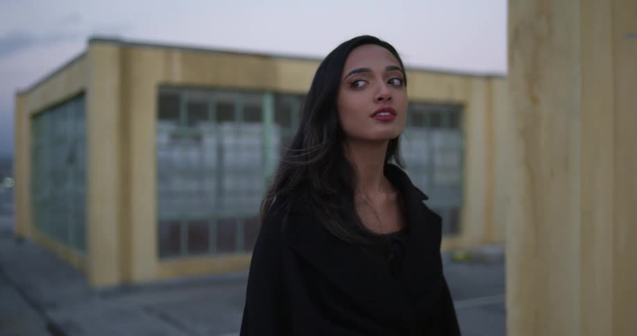 A woman in black walks past two buildings with many windows and looks around