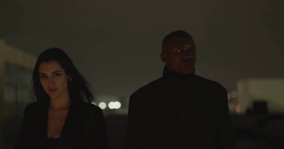Black man and white woman working partners walking together in a subterranean garage in slow motion with headlights crossing them periodically