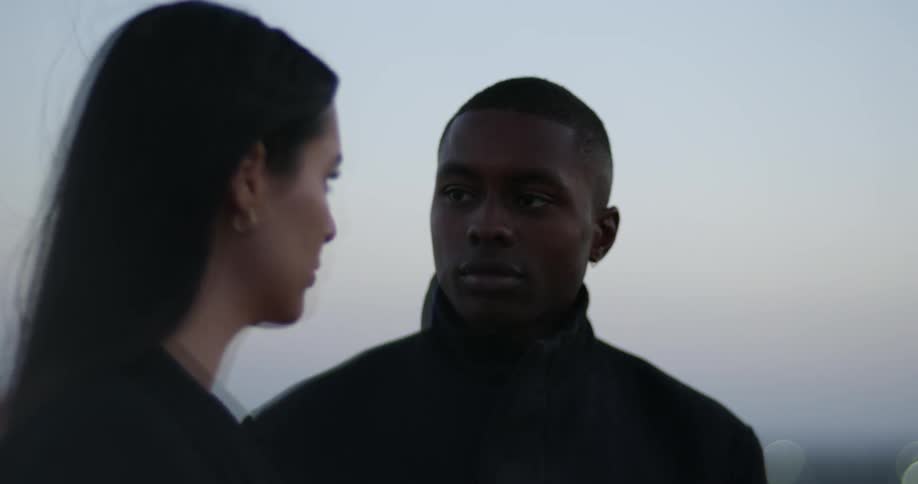 Man with black overcoat stares at woman with dark hair and blazer jacket on top of building at dusk