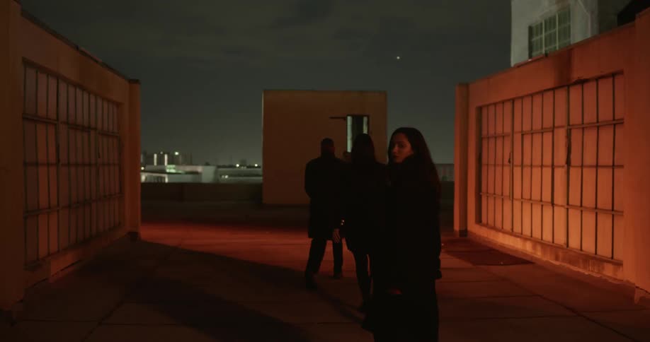 Three people in dark coats walking between building with flashing lights at night