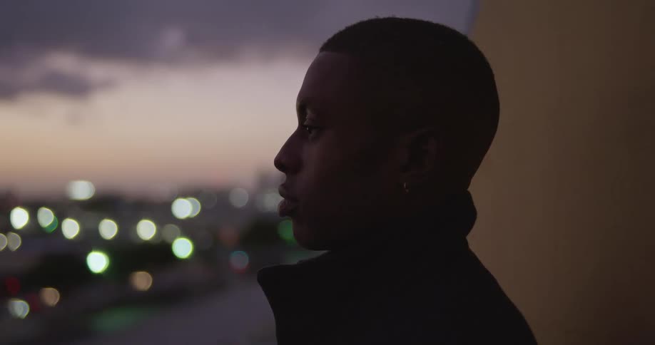 Side profile of shaved head man in black coat looking into space on top of building at dusk