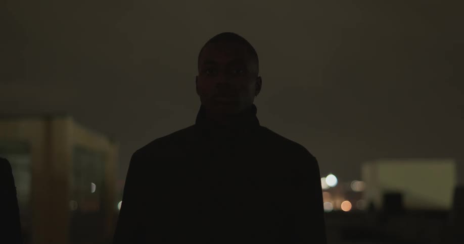 A black man in black clothing walks towards the camera looking forward with a city backdrop lit up at night