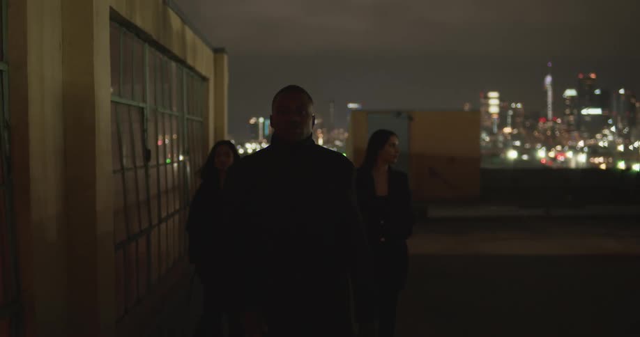 Three people walk across building roof at night with bright city skyline in the background