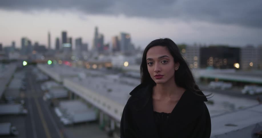 A woman in black stands in front of a city and parked trucks with storm clouds overhead staring at the camera in a portrait fashion