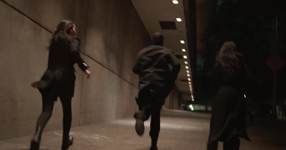 Three people wearing dark coats run alongside building at night