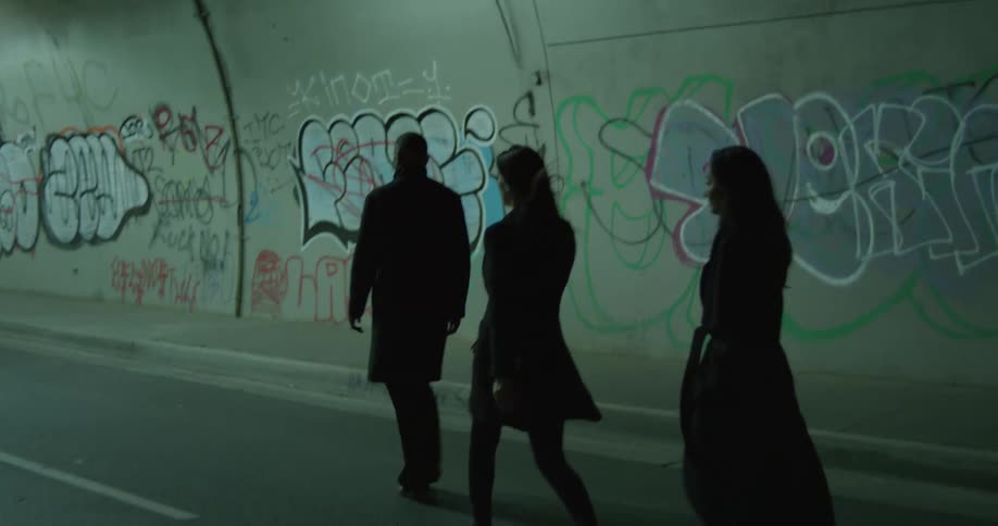 Man in black coat and women wearing black coat and blazer jacket walk through tunnel with graffitis painted on walls