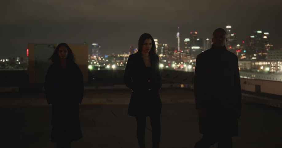 Women in black coat and blazer jacket and man in black overcoat walk on top of building with illuminated skyline in the background at night