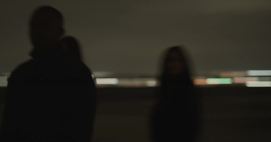 Two white women and a black man chaotic and moving erratically together outside at nighttime hugging stumbling looking around with red lights flashing on them