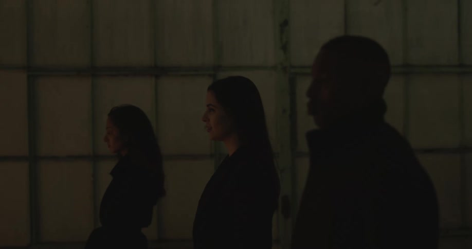Two women and a man all dressed in black are walking in slow motion with a spotlight flashing on them intermittently