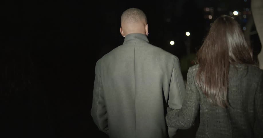 A woman is slightly behind a man whos arm is being held by a woman on the other side of him keeping pace as they all walk briskly down the nighttime downtown sidewalk a spotlight flashes over their backs periodically