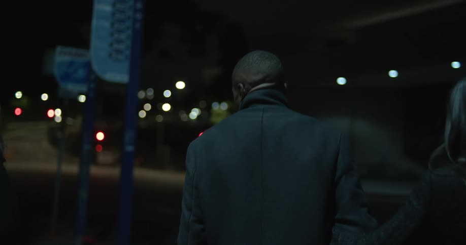 Three people leaning together walk away from building and down street at night with one couple arm in arm