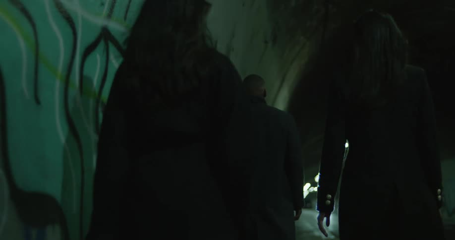 Two women and one man wearing black walk through a tunnel with graffiti on the walls