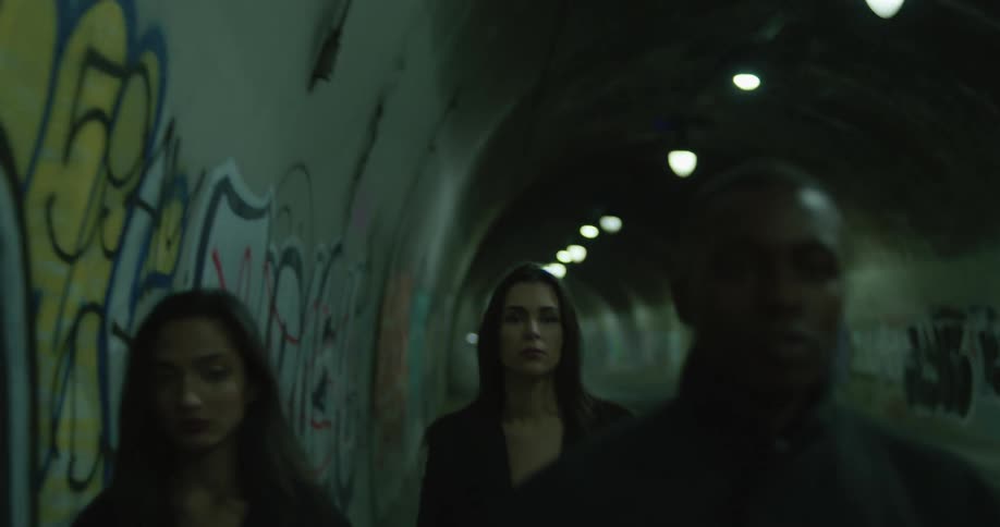 Two women and a man walking together toward cam in a lit up tunnel with graffiti on the walls
