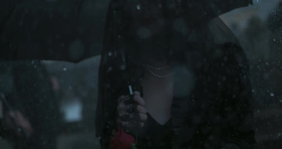 A woman in mourning clothes holds an umbrella and a red rose in the rain
