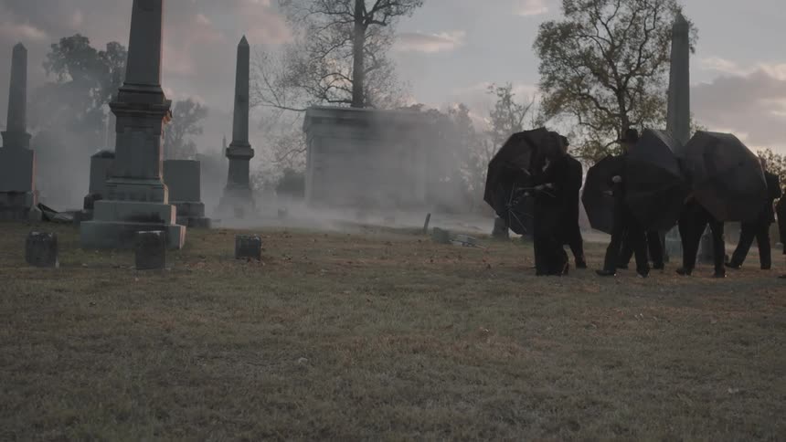 People in black holding umbrellas walk in a cemetery