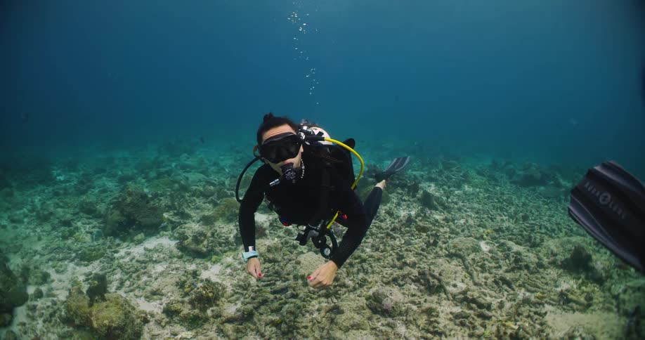 One scuba diver and another mostly offcamera swimming close to coral ocean floor in relatively shallow blue water while looking at other divers flipper and blowing bubbles
