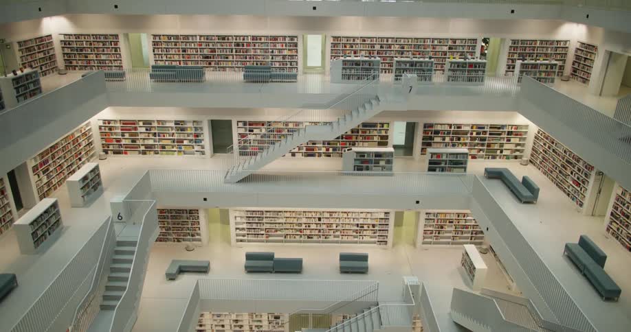 A huge library with many floors and book shelves lining the walls with stairs and furniture on the floors