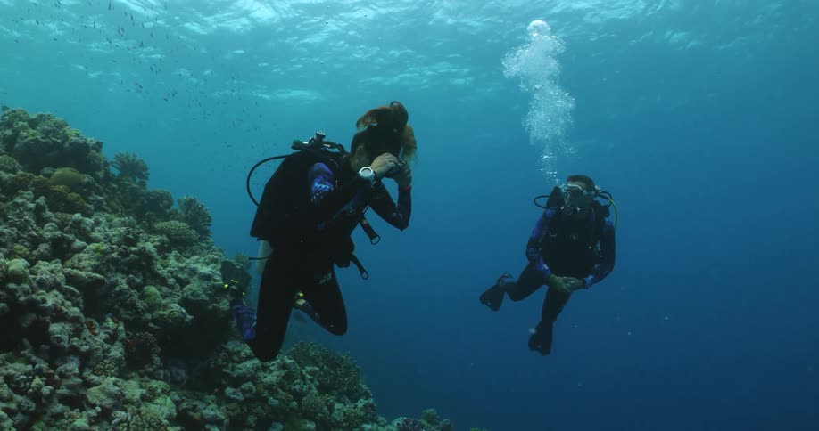 Two scuba divers release bubbles toward the surface while swimming underwater nearreef