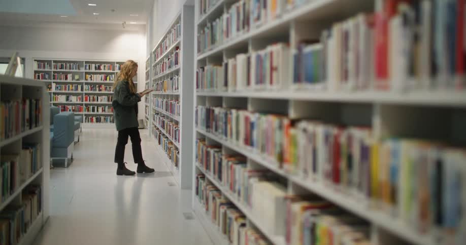 A woman walking along a bookshelf in a library and then stopping to pull a book to look at