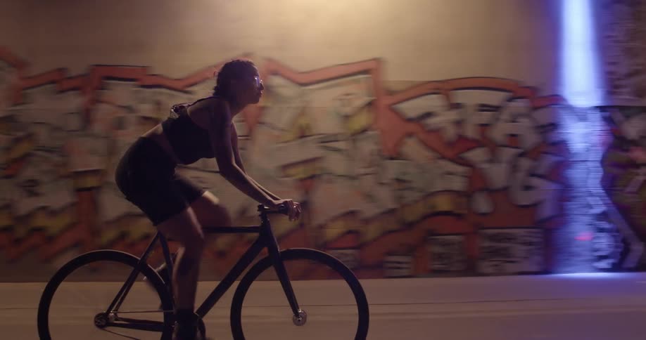 A woman is riding her bicycle on past buildings with pictures painted on the walls on a street at night