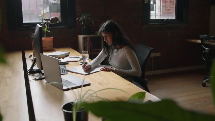 A woman is sitting at a desk in front of computers as she writes on a paper and works on the computer
