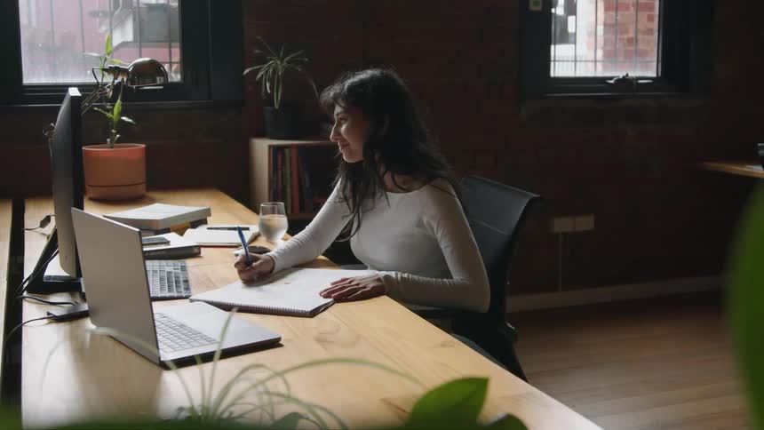 A woman is sitting at a desk in front of computers as she writes on a paper and works on the computer