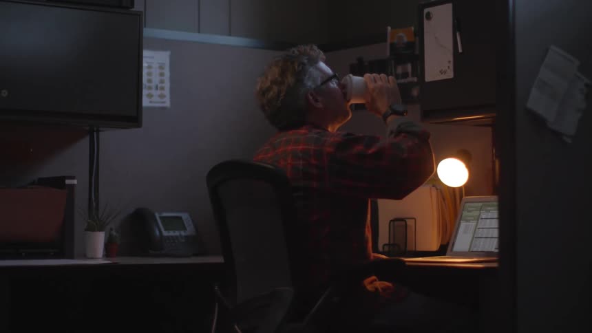 Adult man sitting on a cubicle in the office at night working on a laptop while drinking coffee