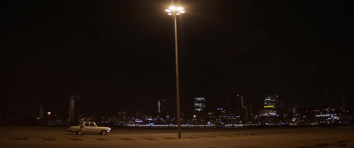 Vintage car parks under a streetlight and a passenger steps out in front of the city skyline at night