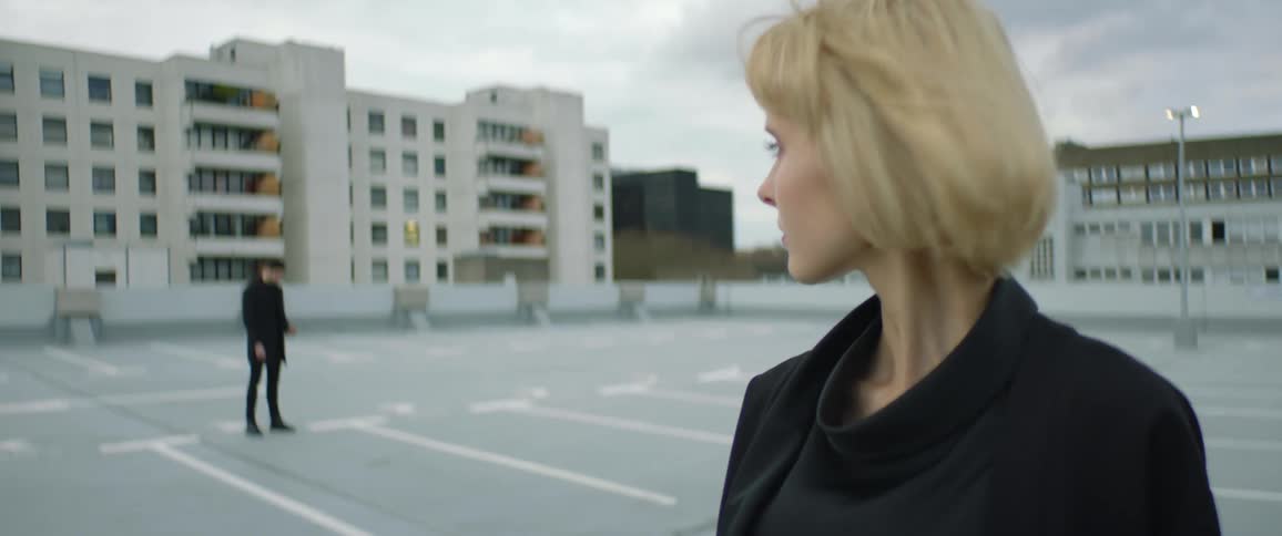 A man stands on a rooftop and looks at his phone and then looks up at a woman across the rooftop who turns and looks at him
