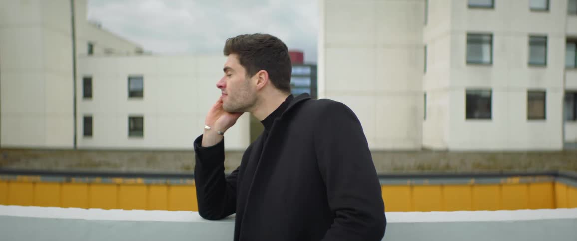 A frustrated man on a rooftop looks around and taps his hand on a railing while he waits