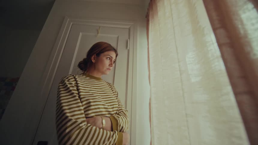A woman is standing near a door as she looks out a window on the side of a curtain