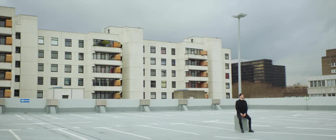 Man sits at base of parking lot light in empty parking lot near apartment buildings