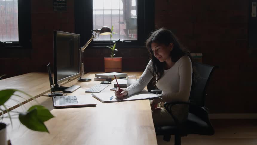 A woman sitting at her desk using a pencil to write on paper in front of a computer screen