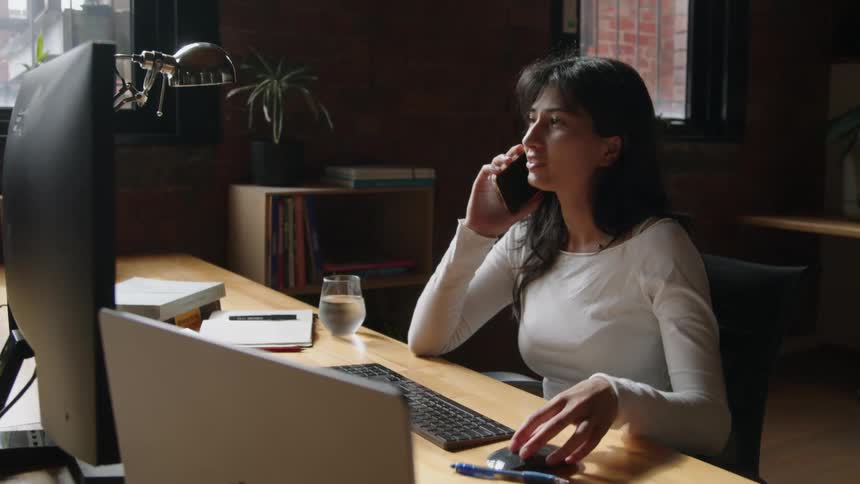 A woman sits at a desk in an office while talking on the phone and working on a computer