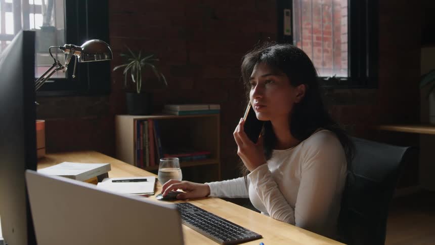Young woman sitting at a desk in the office speaks on her phone while working on a computer screen