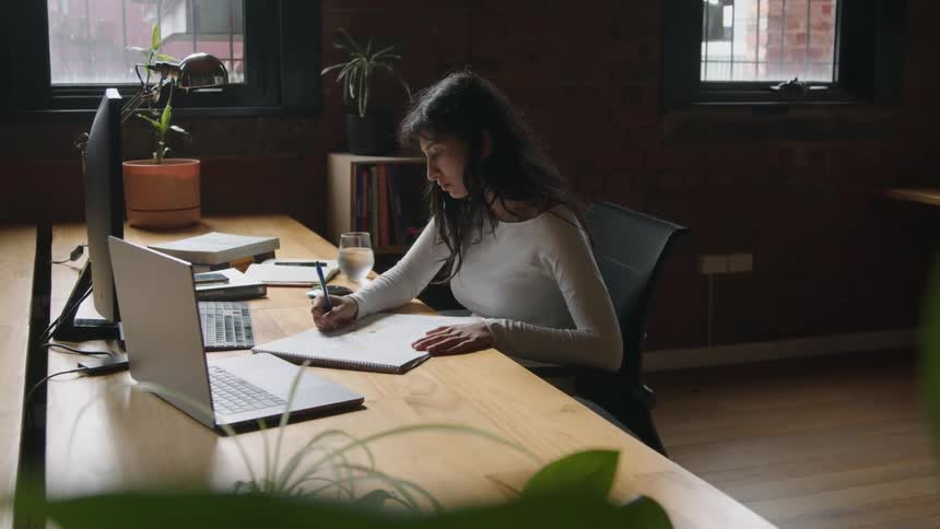 Young woman sitting at a desk in the office writes notes while reading on a computer screen