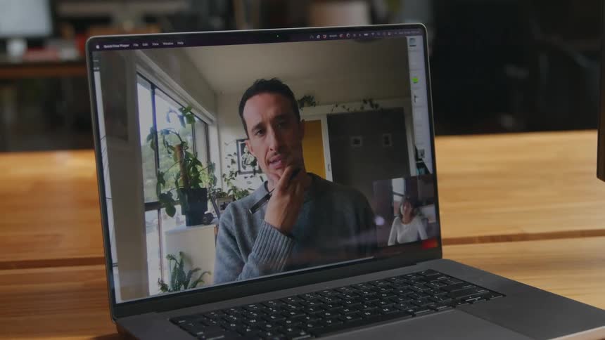 Laptop sitting on wooden table shows video of man in office talking