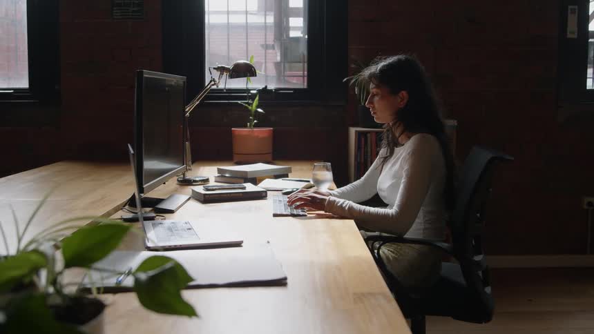 A woman sits at a long table typing on a computer in an office