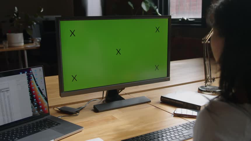 Woman sits working at desk with laptop and large computer monitor