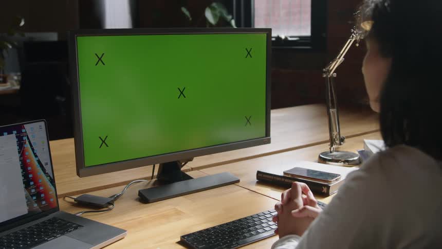 Young woman sitting at a desk in the office looking at a monitor with a green replaceable screen